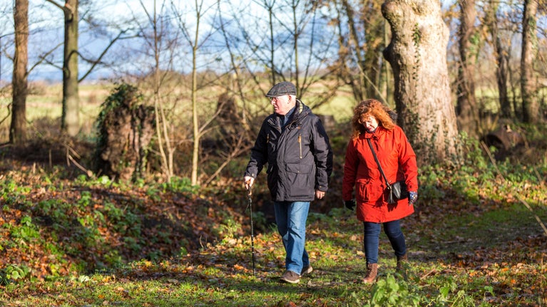 A man and woman walking through the woodland at Rufford Old Hall in winter. There is a low sun behind them.